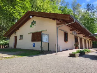 Ein einfaches Gebäude umgeben von grünen Bäumen. Der Eingang ist gepflastert und einige Pflanzen sind vor der Fassade.