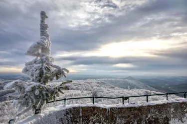Ein verschneiter Aussichtspunkt mit Blick auf eine winterliche Landschaft. Im Hintergrund sind schneebedeckte Hügel und ein bewölkter Himmel zu sehen.