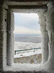 Ein verschneiter Ausblick durch ein gefrorenes Fenster. Die Landschaft ist von Eis und Schnee bedeckt, und der Himmel ist bewölkt.