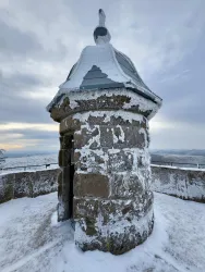 Ein schneebedeckter Aussichtsturm mit einer steinernen Struktur. Der Himmel ist bewölkt und es liegt eine winterliche Landschaft um den Turm herum.