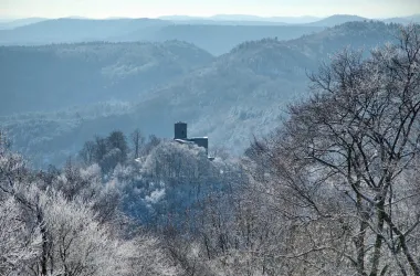 Eine schneebedeckte Landschaft mit hügeligen Bergen im Hintergrund. Auf einem Hügel steht eine alte Burg, umgeben von frostigen Bäumen.