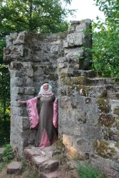 Eine Frau in historischer Kleidung steht an einer alten Steinmauer. Die Umgebung ist grün und sonnig.