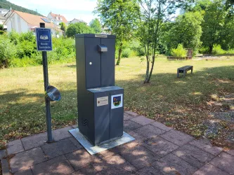 Ein Trinkbrunnen steht in einem parkähnlichen Bereich. Im Hintergrund sind Bäume und eine Bank zu sehen.