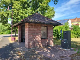 Eine kleine Bushaltestelle aus Stein mit einem Satteldach steht an einer grünen Straße. Neben der Haltestelle befindet sich ein Trinkbrunnen.