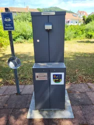 Eine Trinkwasserstation mit einem grauen Gehäuse steht in einem grünen Bereich. Daneben befindet sich ein Hinweiszeichen für die Wasserentnahme.