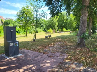 Ein ruhiger Park mit grünen Bäumen und einer Landschaft aus Gras. Im Vordergrund steht ein Trinkbrunnen neben einer Bank.