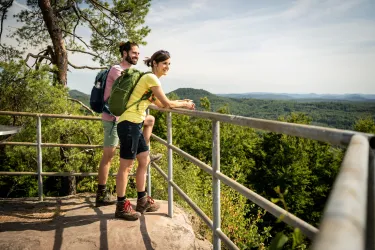 Ein Paar genießt die Aussicht von einem Aussichtspunkt in der Natur. Im Hintergrund sind grüne Hügel und ein blauer Himmel zu sehen.
