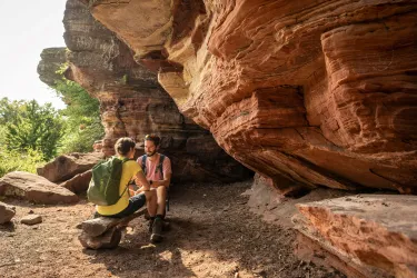 Zwei Personen sitzen unter einer großen Felsformation und unterhalten sich. Die Umgebung besteht aus rotem Fels und grünen Bäumen im Hintergrund.
