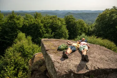 Ein Paar liegt entspannt auf einem großen Felsen und genießt die Aussicht auf die grüne Landschaft. Im Hintergrund sind sanfte Hügel und Wälder zu sehen.