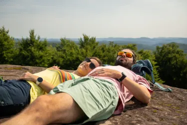 Ein Paar liegt entspannt auf einem Felsen und genießt die Aussicht. Im Hintergrund sind Wälder und sanfte Hügel zu sehen.