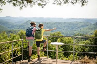 Zwei Wanderer stehen auf einem Aussichtspunkt und blicken auf die bewaldeten Hügel. Die Sonne scheint und die Landschaft ist grün und einladend.