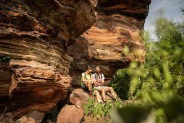 Ein Paar sitzt entspannt unter einem großen Felsen und genießt einen Moment in der Natur. Die Umgebung zeigt beeindruckende steinerne Formationen und grünes Laub.
