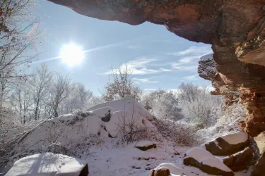 Ein winterlicher Landschaftsausblick durch eine Felsenöffnung. Der Boden ist mit Schnee bedeckt und die Sonne scheint hell am Himmel.