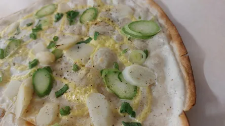 Eine dünne Pizza mit grünen Zwiebeln, weißen Spargelstücken und einer cremigen Sauce. Die Oberfläche ist leicht gewürzt und wirkt frisch.