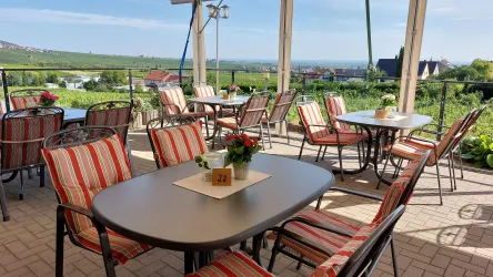 Ein einladender Außenbereich mit Tischen und Stühlen, umgeben von Weinbergen. Die Sonne scheint und bietet eine entspannte Atmosphäre.