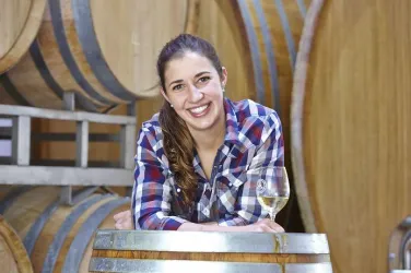 Eine lächelnde Frau steht an einem Weinfass und hält ein Glas Wein in der Hand. Im Hintergrund sind weitere Fässer zu sehen.