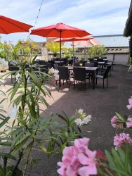 Eine Terrasse mit schwarzen Tischen und Stühlen, die von roten Sonnenschirmen beschattet wird. Bunte Blumen umgeben den Bereich und verleihen eine angenehme Atmosphäre.