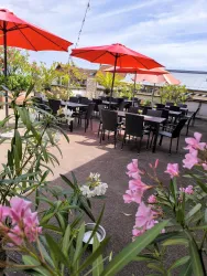 Eine gemütliche Terrasse mit schwarzen Tischen und roten Sonnenschirmen. Umgeben von bunten Pflanzen und einem klaren Himmel.