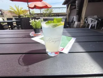 Ein erfrischender Cocktail mit Minze und Zitrone steht auf einem Tisch. Im Hintergrund sind sonnige Stühle und eine Terrasse zu sehen.