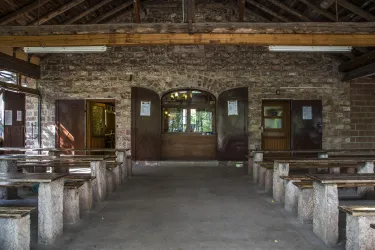 Ein rustikaler Innenraum mit Steinmauern und Holzbänken. Auf der Vorderseite befinden sich große Fenster und Türen, die den Blick nach draußen bieten.
