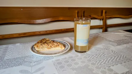Ein Stück Kuchen auf einem Teller und ein Glas mit einem Getränk stehen auf einem Tisch. Der Tisch ist mit einer hellen Tischdecke gedeckt.