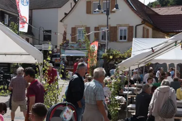Ein belebter Marktplatz mit mehreren Ständen und vielen Menschen. Im Hintergrund sind traditionelle Gebäude zu sehen, umgeben von Festzeltstrukturen.