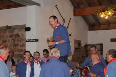 Eine Gruppe von Männern in blauen Hemden feiert in einem rustikalen Raum. Ein Mann steht auf einem Tisch und hält ein großes Glas Bier.