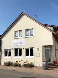Ein freundliches Weingut mit einem einladenden Eingang und bunten Dekorationen. Die Fassade ist hell und freundlich, umgeben von grünen Pflanzen.