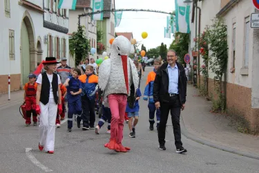 Ein bunter Umzug mit mehreren Gruppen von Menschen auf der Straße. Eine Person trägt ein auffälliges Kostüm und ein Bürgermeister lächelt in der Menge.
