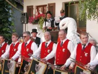 Eine Gruppe von Musikern in roten Westen spielt vor einem festlichen Hintergrund. Im Hintergrund steht eine Person mit einem Kostüm und einer Figur eines Schwans.