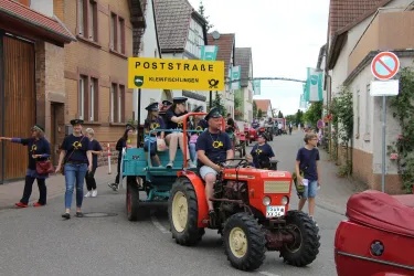 Ein kleiner Umzug mit einem Traktor, der eine bunte Gruppe von Personen zieht. Die Teilnehmer tragen T-Shirts und feiern in einer Straße mit Häusern im Hintergrund.