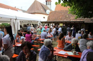 Eine lebhafte Veranstaltung im Freien mit vielen Menschen, die an Tischen sitzen. Im Hintergrund sind Zelte und ein Gebäude mit einem traditionellen Dach zu sehen.