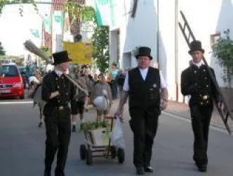Eine Gruppe von Männern in historischen Kostümen marschiert eine Straße entlang. Sie tragen Besen und einen Wagen mit Tieren.