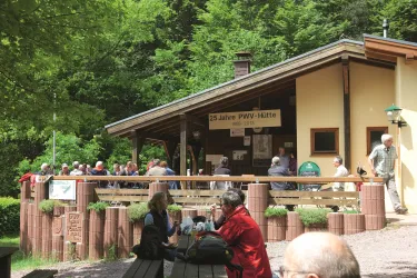Eine Hütte im Wald mit einer Gruppe von Menschen, die sich versammelt haben. Die Umgebung ist grün und einladend.