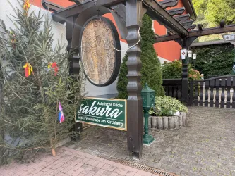 Ein Eingangsbereich mit einem großen Holzportal und einem Schild, das asiatische Küche bewirbt. Links steht ein Tannenbaum mit bunten Anhängern.