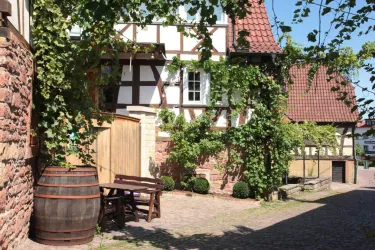 Ein malerischer Innenhof mit Weinranken und einer alten Holzfass. Im Hintergrund sind Fachwerkhäuser und eine gemütliche Sitzgelegenheit zu sehen.