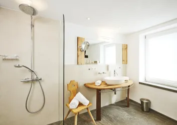 Ein modernes Badezimmer mit einer ebenerdigen Dusche und einem kleinen Holzstuhl. Der Raum ist hell und freundlich mit Fenstern, die viel Licht hereinlassen.