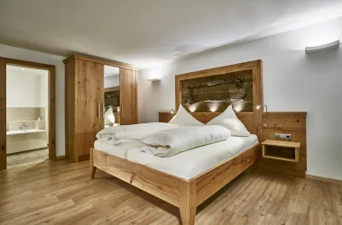 Ein modernes Schlafzimmer mit einem Holzbett und gemütlicher Bettwäsche. Im Hintergrund sieht man einen Schrank und einen Zugang zum Badezimmer.