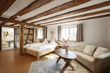 Ein modernes Zimmer mit Holzbalkendecke, einem großen Bett und einem gemütlichen Sofa. Besonders hervorzuhebende Details sind der Teppich und die Fenster mit viel Tageslicht.