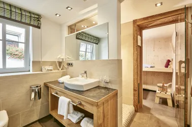 Ein modernes Badezimmer mit hellem Design und Holzdetails. Im Hintergrund ist eine Sauna erkennbar.