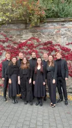 Eine Gruppe von zehn Personen steht vor einer steinernen Wand mit roten Blättern. Alle tragen schwarze Kleidung und lächeln in die Kamera.