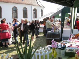 Ein belebter Markt mit Ständen und Besuchern. Im Vordergrund sind Produkte und Blumen zu sehen.