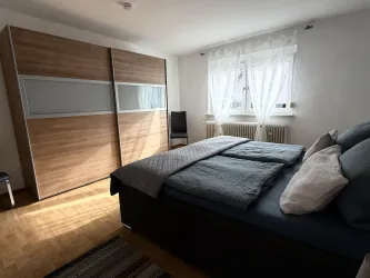 Ein helles Schlafzimmer mit einem großen Bett und einer modernen Holzschrankwand. Die Fenster lassen viel Tageslicht rein und es gibt einen Stuhl in der Ecke.
