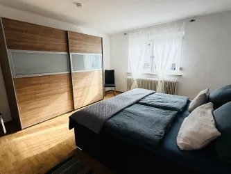 Ein helles Schlafzimmer mit einem großen Bett und einer modernen Holzschrankwand. Durch das Fenster fällt sanftes Licht in den Raum.
