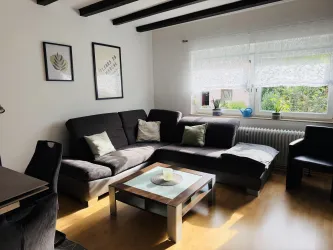 Ein gemütliches Wohnzimmer mit einer dunklen Couch und einem Tisch in der Mitte. Große Fenster lassen viel Licht herein und es gibt Pflanzen auf der Fensterbank.
