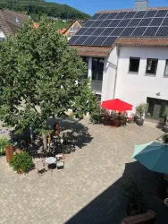 Ein ruhiger Innenhof mit einem großen Baum und Sitzgelegenheiten. Auf der rechten Seite stehen verschiedene Schirme und eine Sonnenliege.