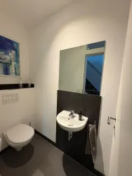 Ein modernes Badezimmer mit einem Handwaschbecken und einem großen Spiegel. An der Wand hängt ein Handtuch und im Hintergrund sind Treppen sichtbar.