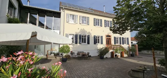 Ein elegantes Haus mit weißer Fassade und blauen Fensterläden. Der Außenbereich ist schön gestaltet mit Blumen und einer Sitzgelegenheit.
