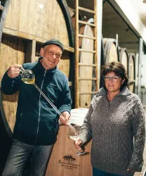 Ein Mann und eine Frau stehen in einem Weinkeller zwischen großen Holzfässern. Der Mann hält ein Glas und eine Glasröhre, während die Frau lächelnd schaut.
