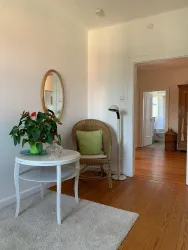 Ein gemütlicher Eingangsbereich mit einem Stuhl, einem kleinen Tisch und einer Pflanze. Der Raum ist hell und freundlich, mit warmen Holzdielen und einem Spiegel an der Wand.